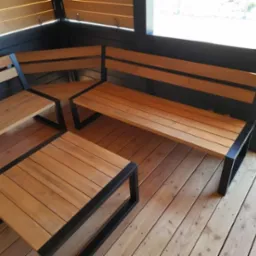 Drewniany zestaw mebli ogrodowych z metalowymi ramami, składający się z narożnej ławki i stolika kawowego, umieszczony na drewnianym tarasie w zacisznym kącie.