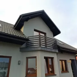 Nowoczesny dom jednorodzinny z szarą elewacją, ciemnobrązową stolarką okienną i balkonem z poziomymi, metalowymi balustradami w kolorze ciemnego drewna.