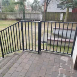 Czarne, metalowe barierki schodowe z pionowymi szczeblami, prowadzące do drzwi wejściowych, z kostki brukowej na podłożu, widok na trawnik i ogrodzenie w tle.