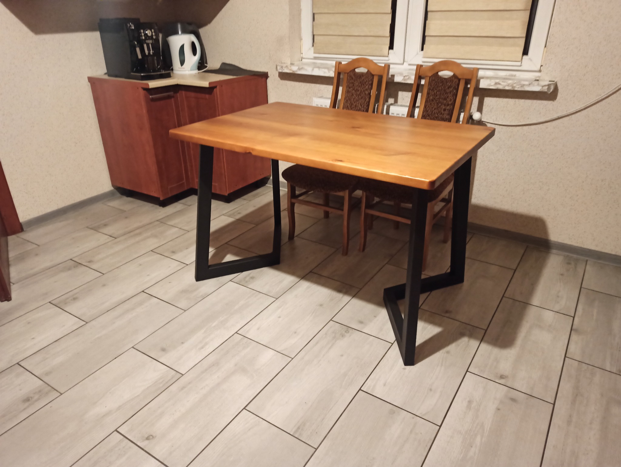 Stół z drewnianym blatem i czarnymi, metalowymi nogami o nietypowym kształcie, ustawiony w pomieszczeniu z jasną podłogą imitującą drewno i dwoma krzesłami w tle.