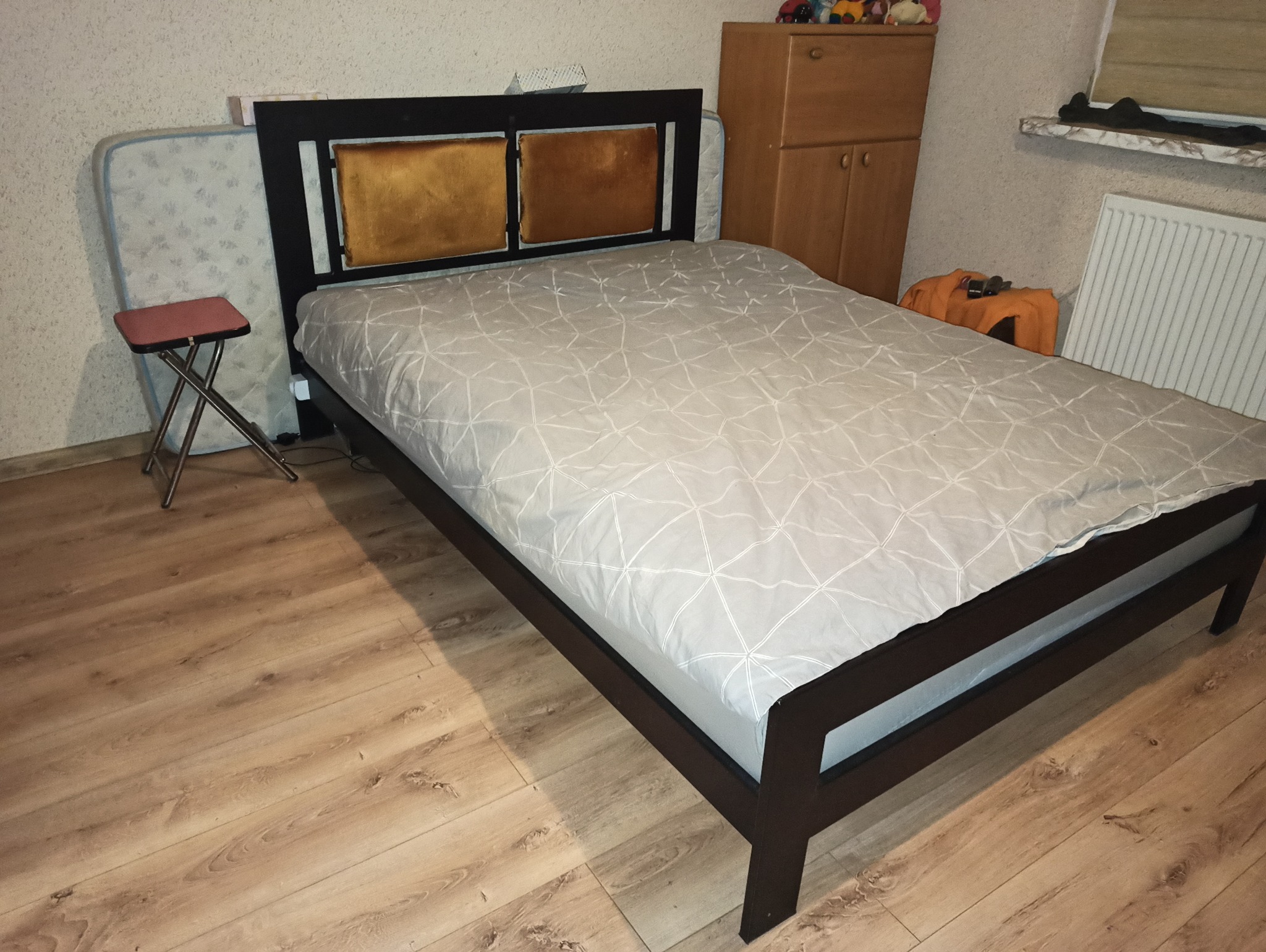 Ciemnobrązowe łóżko z dwoma kwadratowymi zagłówkami w kolorze musztardowym, przykryte białą pościelą w geometryczny wzór, na drewnianej podłodze, z taboretem i materacem opartym o ścianę w tle.