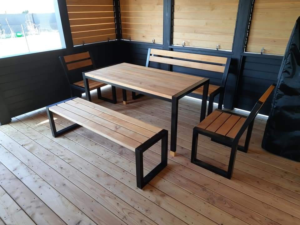 Komplet mebli ogrodowych: stół z drewnianym blatem i czarną metalową ramą, dwie ławki z drewnianymi siedziskami i czarnymi metalowymi nogami oraz dwie krzesła z drewnianymi siedziskami i oparciami...