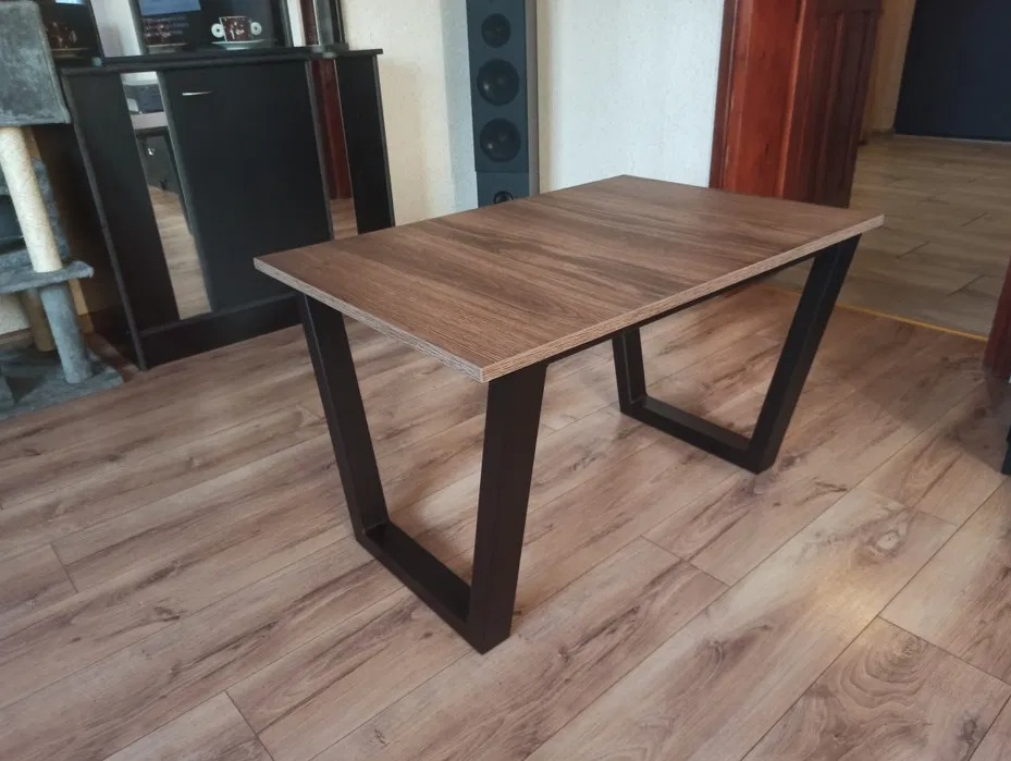 Stół z blatem imitującym drewno i czarnymi, metalowymi nogami w kształcie litery V, ustawiony na panelach podłogowych w pomieszczeniu z meblami.