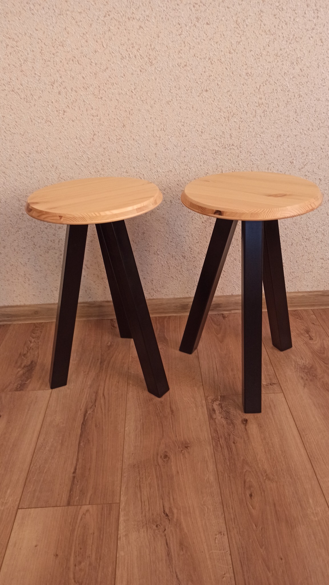 Dwa drewniane taborety z okrągłymi siedziskami i czarnymi, kwadratowymi nogami, ustawione na panelach podłogowych w pomieszczeniu o beżowych ścianach.