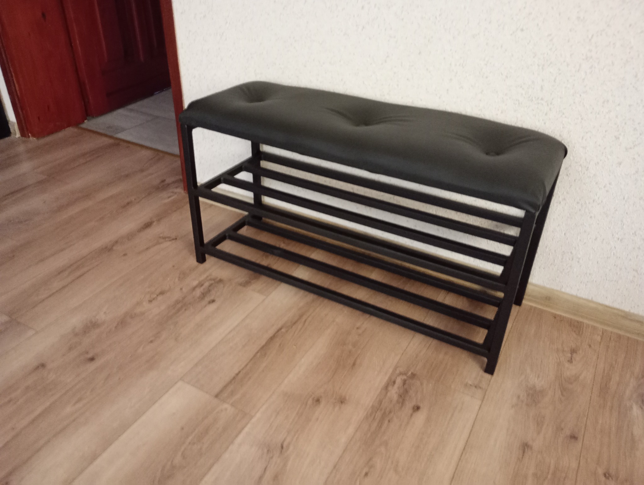 Czarna metalowa ławka z tapicerowanym siedziskiem i półkami na buty, ustawiona na panelach podłogowych przy białej ścianie.