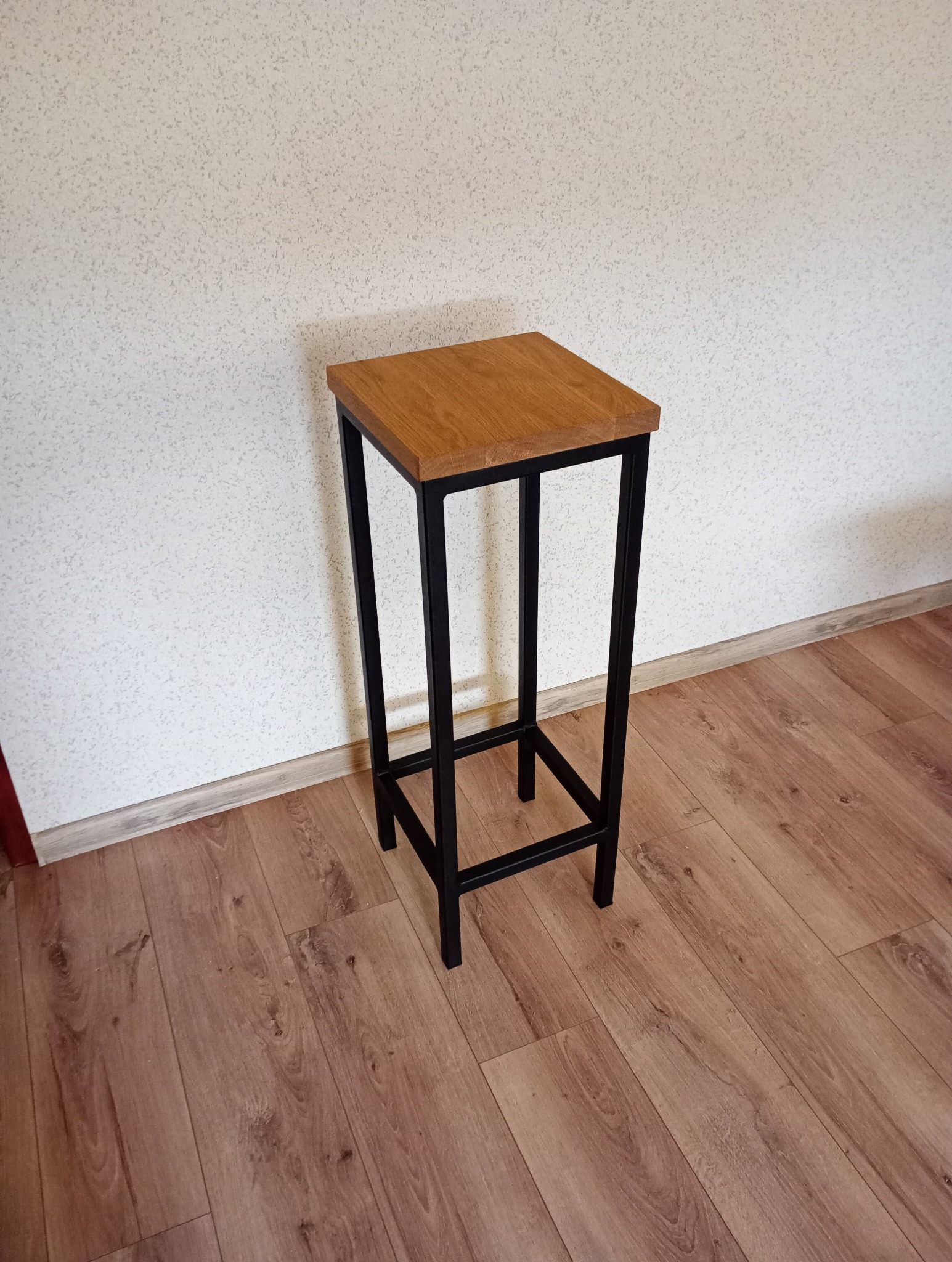 Wysoki stołek barowy z drewnianym siedziskiem i czarną, metalową ramą, ustawiony na panelach podłogowych w pomieszczeniu z jasną ścianą o drobnej fakturze.