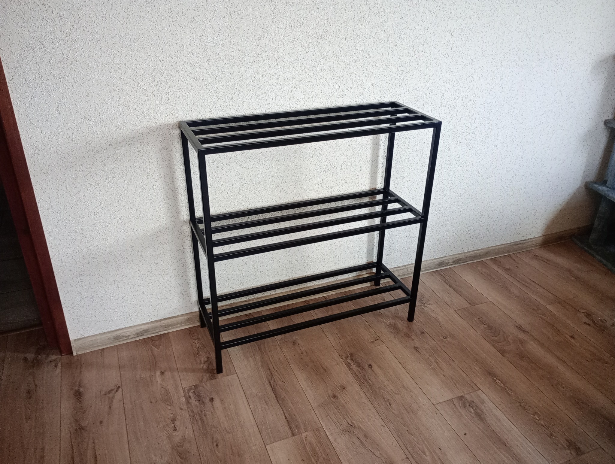 Trzypoziomowy, czarny metalowy regał na buty z prostokątnymi szczeblami, stojący na drewnianej podłodze przy białej ścianie o nierównej fakturze.
