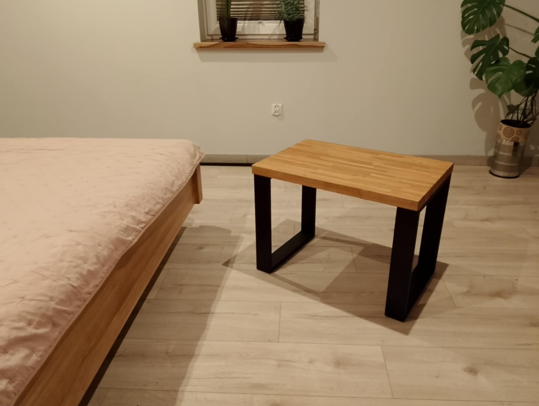 Stolik kawowy z blatem z jasnego drewna i czarnymi, metalowymi nogami w nowoczesnym wnętrzu sypialni, obok łóżka z różową narzutą i dużej rośliny doniczkowej.