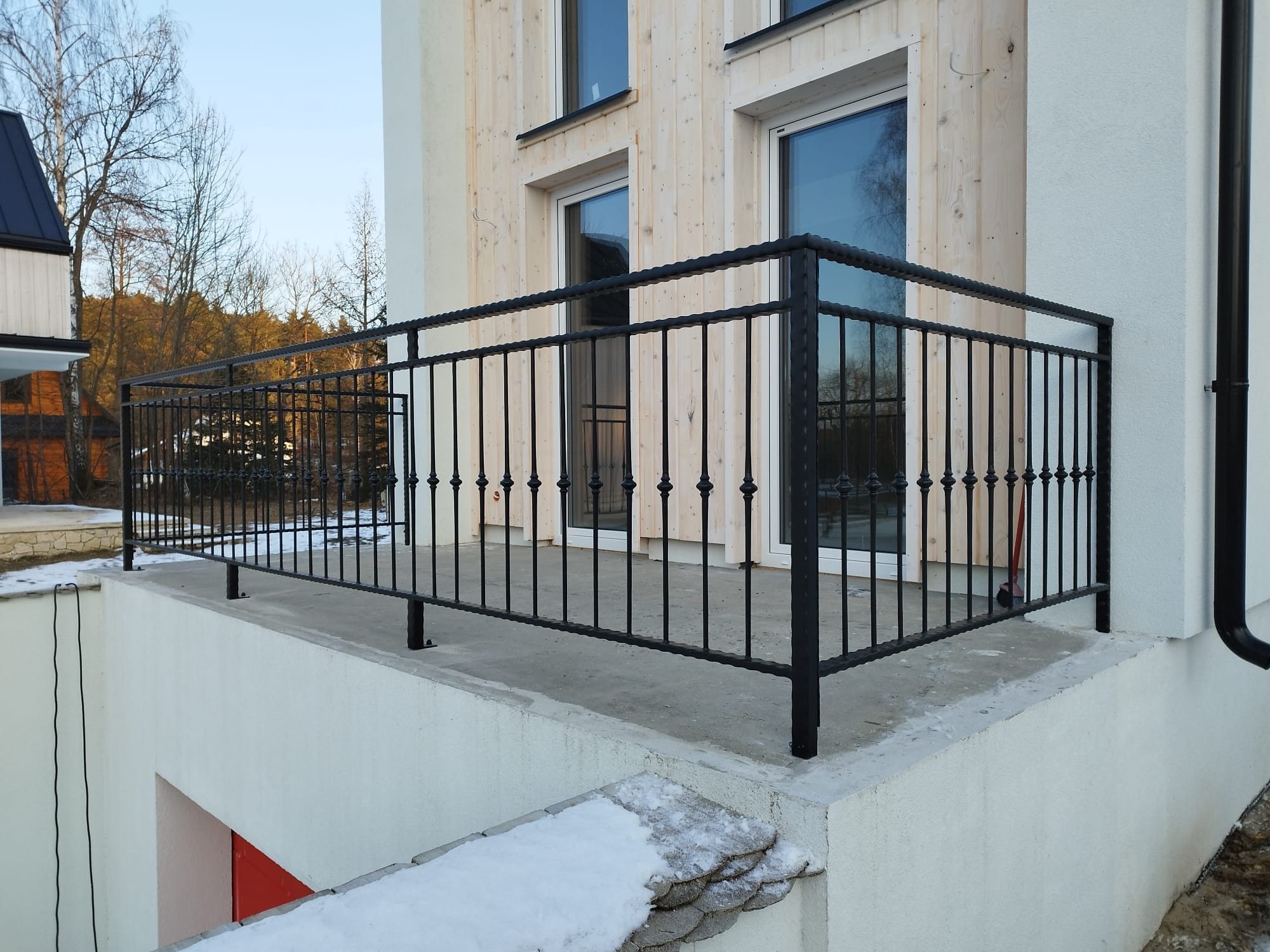 Czarna, metalowa balustrada balkonowa z dekoracyjnymi elementami na tle jasnej elewacji budynku z drewnianymi panelami i widocznym fragmentem zaśnieżonego terenu.