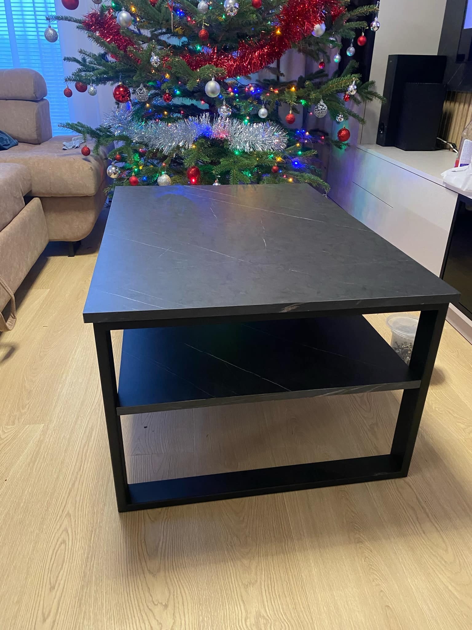 Stolik kawowy z czarnym metalowym stelażem i blatem imitującym kamień, z dodatkową półką, ustawiony w salonie z choinką w tle.