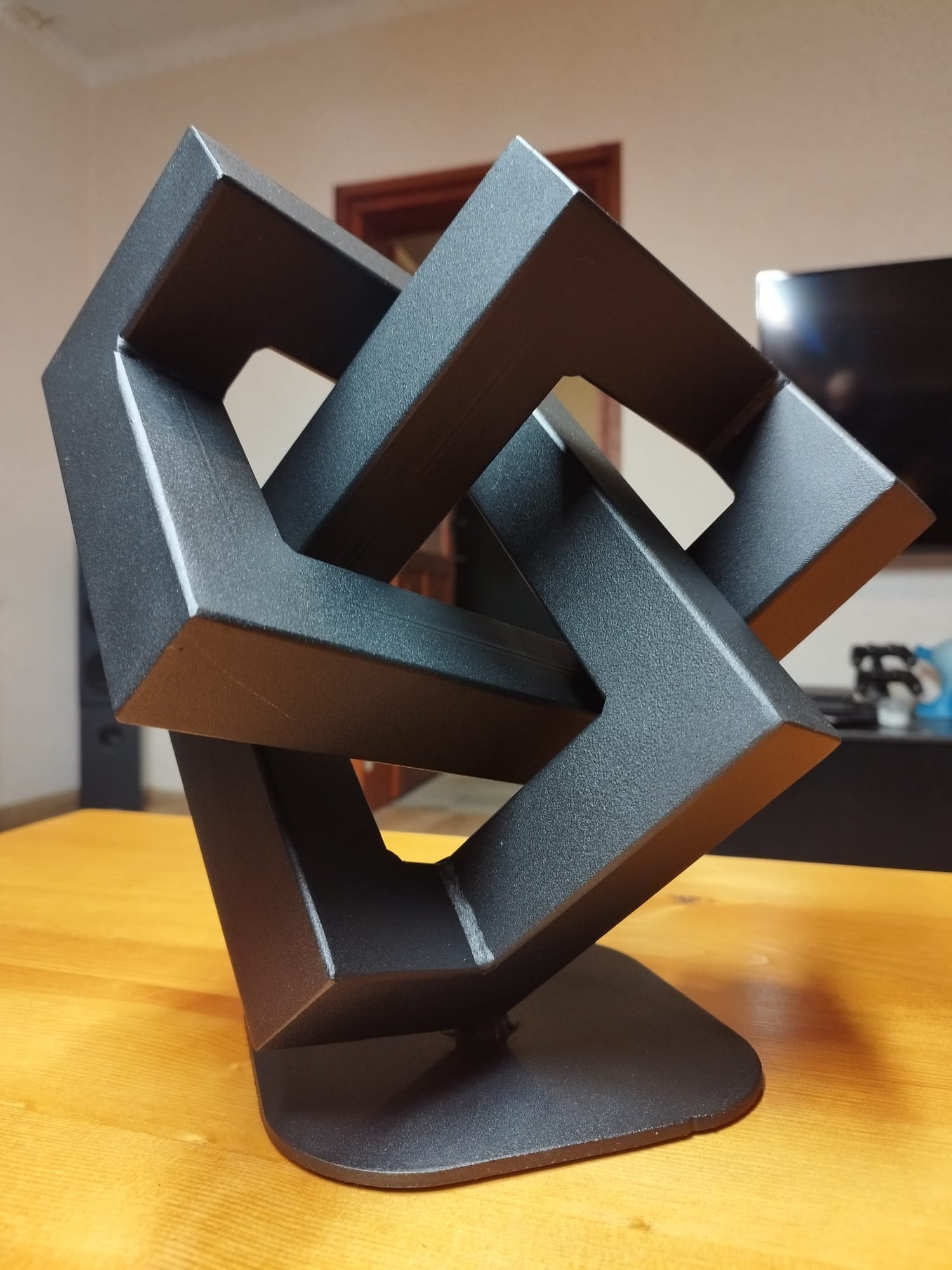 Czarna, metalowa rzeźba przestrzenna o geometrycznym kształcie, stojąca na drewnianym blacie, z widoczną teksturą powierzchni i subtelnymi refleksami światła.