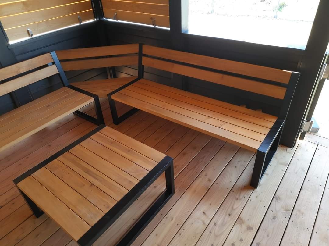 Drewniany zestaw mebli ogrodowych z metalowymi ramami, składający się z narożnej ławki i stolika kawowego, umieszczony na drewnianym tarasie w zacisznym kącie.