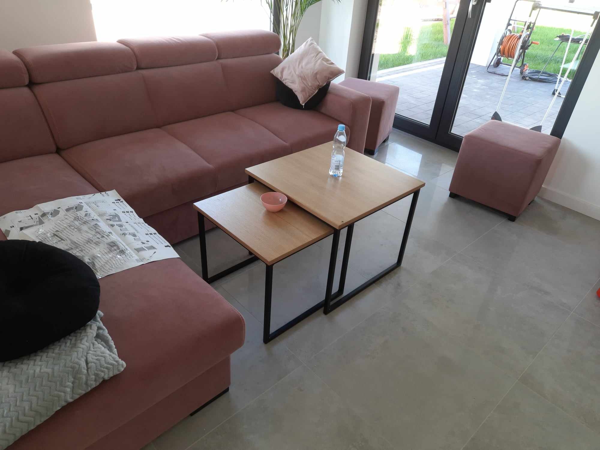 Dwa stoliki kawowe z drewnianymi blatami i czarnymi, metalowymi nogami w nowoczesnym salonie z narożną kanapą w odcieniach różu, butelką wody i miseczką na blacie, na podłodze szare płytki.