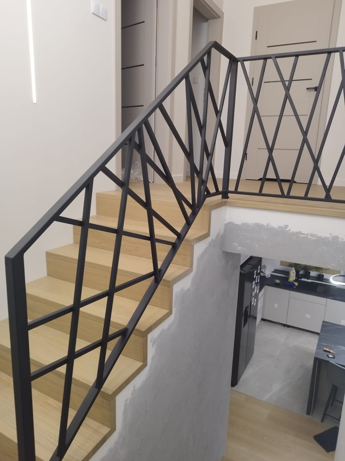 Nowoczesne poręcze schodowe z metalową konstrukcją w geometryczny wzór, widok na jasne drewniane schody i fragment kuchni w Starym Majdanie.