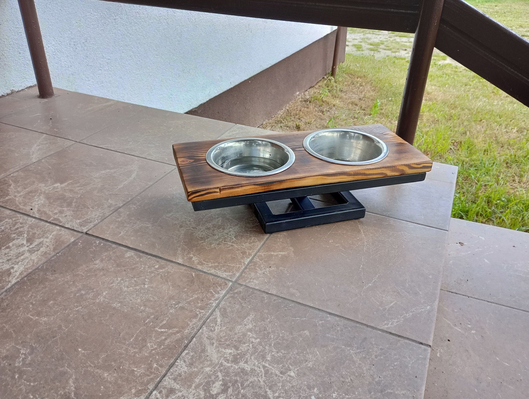 Podwyższony stojak na dwie miski dla zwierząt, wykonany z ciemnego, bejcowanego drewna i czarnej metalowej podstawy, umieszczony na schodach wyłożonych płytkami.