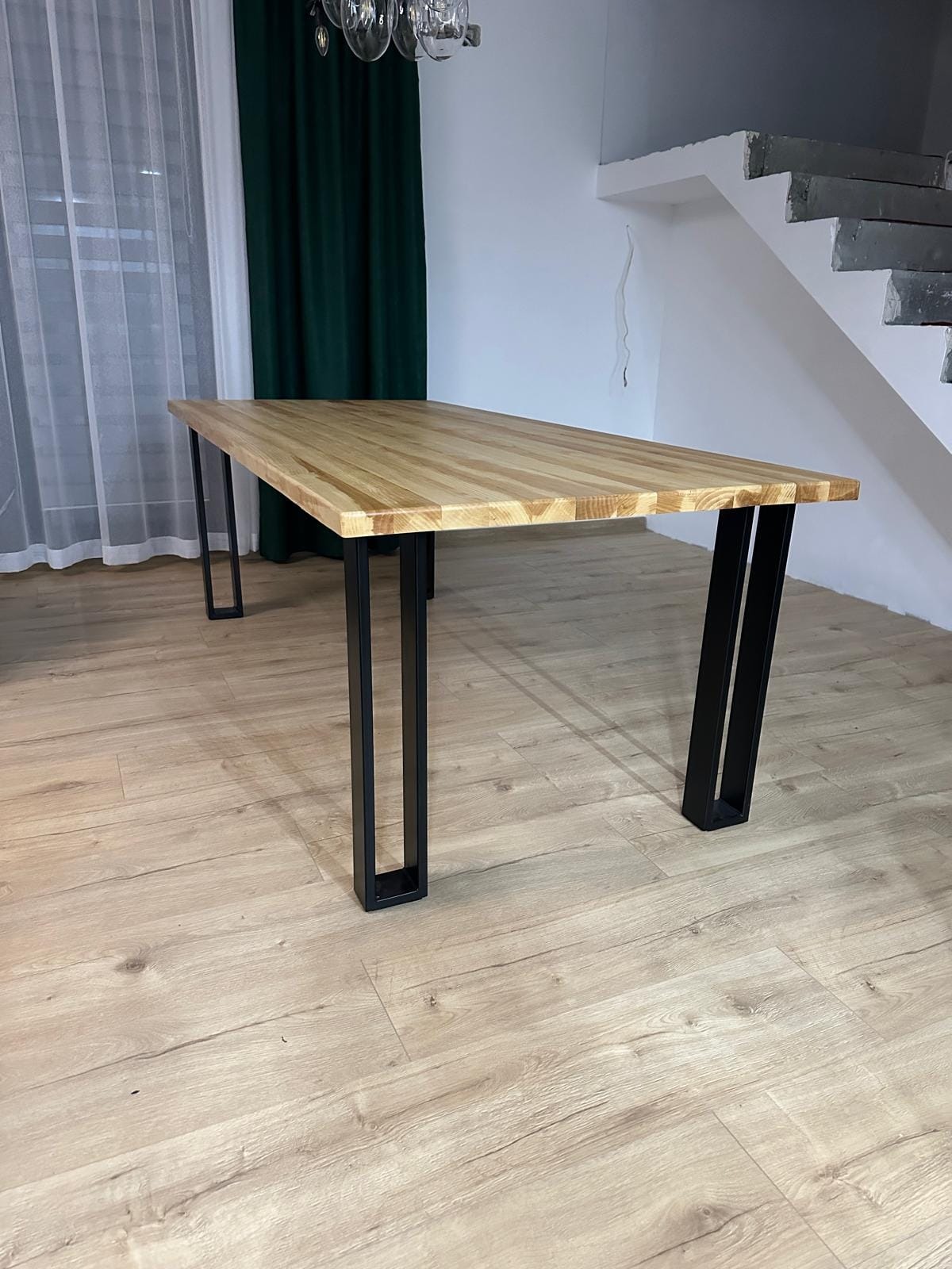Stół z blatem z jasnego drewna i czarnymi, metalowymi nogami o prostokątnym przekroju, na tle zasłon i schodów.