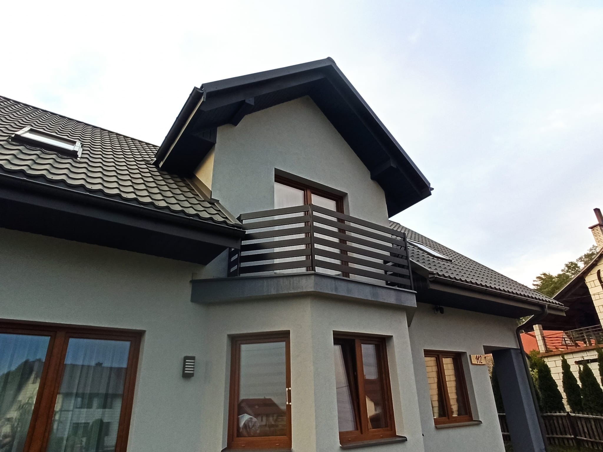 Nowoczesny dom jednorodzinny z szarą elewacją, ciemnobrązową stolarką okienną i balkonem z poziomymi, metalowymi balustradami w kolorze ciemnego drewna.