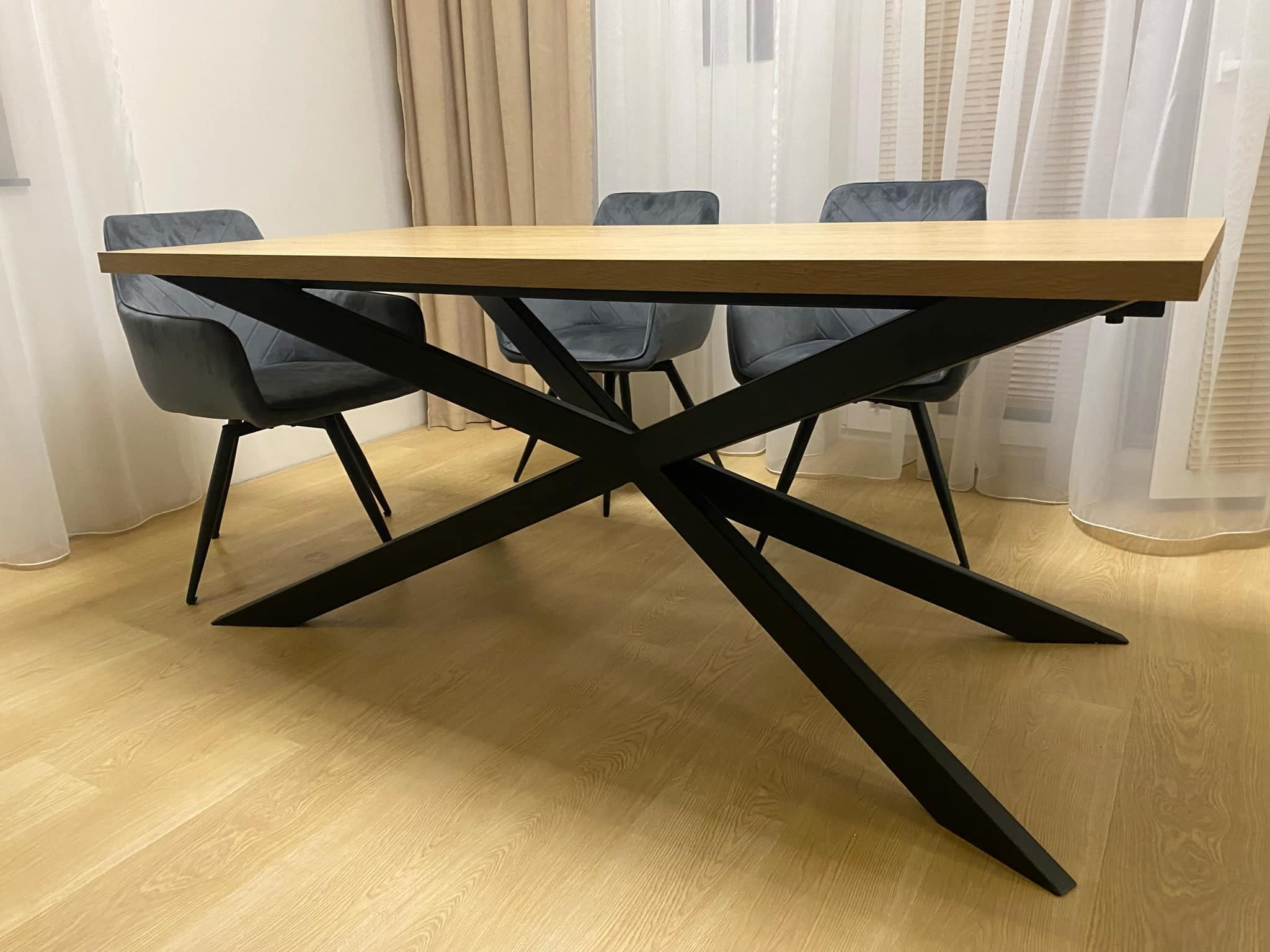 Stół z jasnym blatem i czarną, metalową podstawą w kształcie litery X, otoczony trzema szarymi krzesłami na tle okna z białymi zasłonami.
