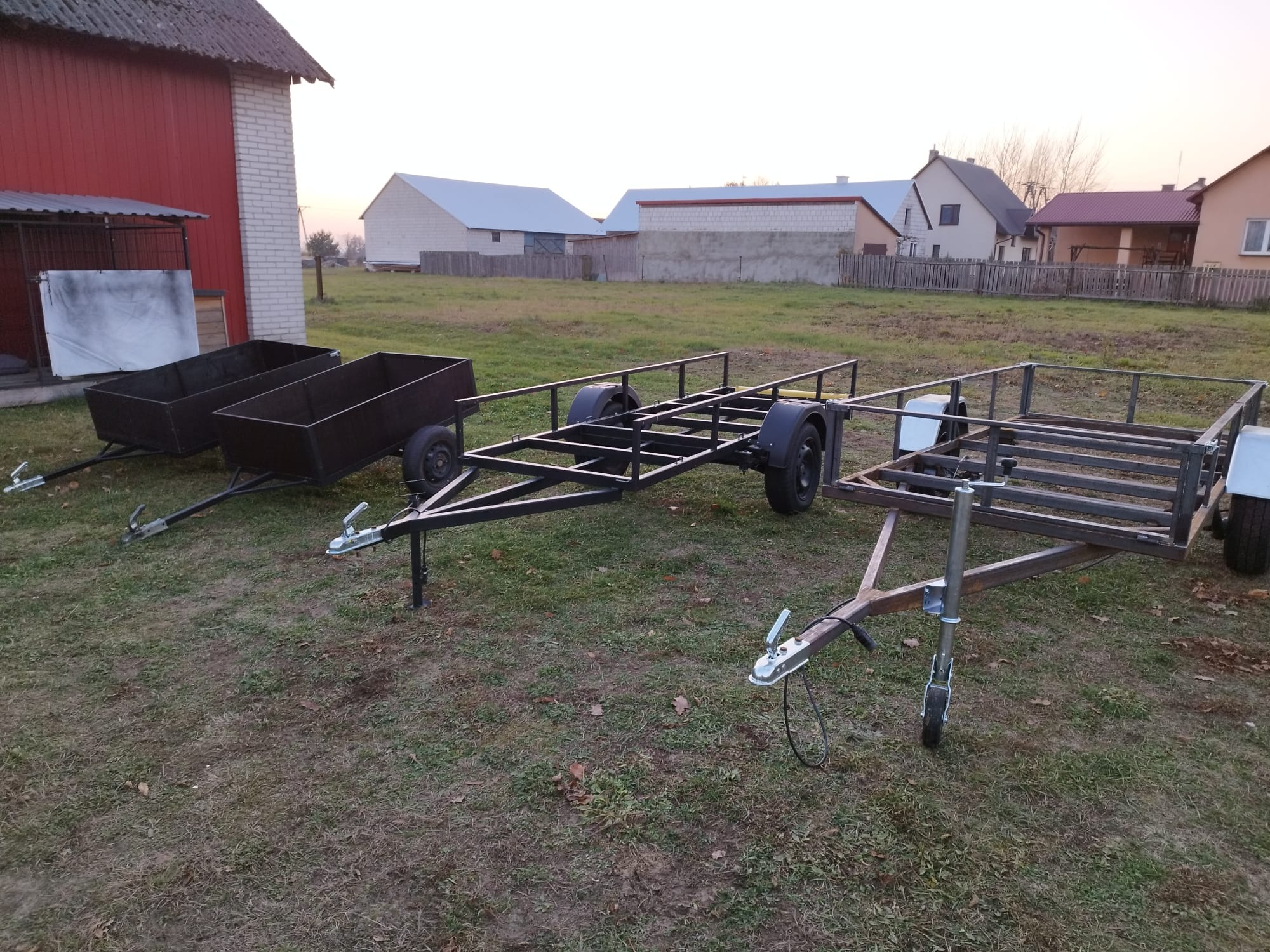 Trzy przyczepki na trawie: dwie z metalowymi skrzyniami, jedna z ramą z profili, w tle budynki mieszkalne i gospodarcze.