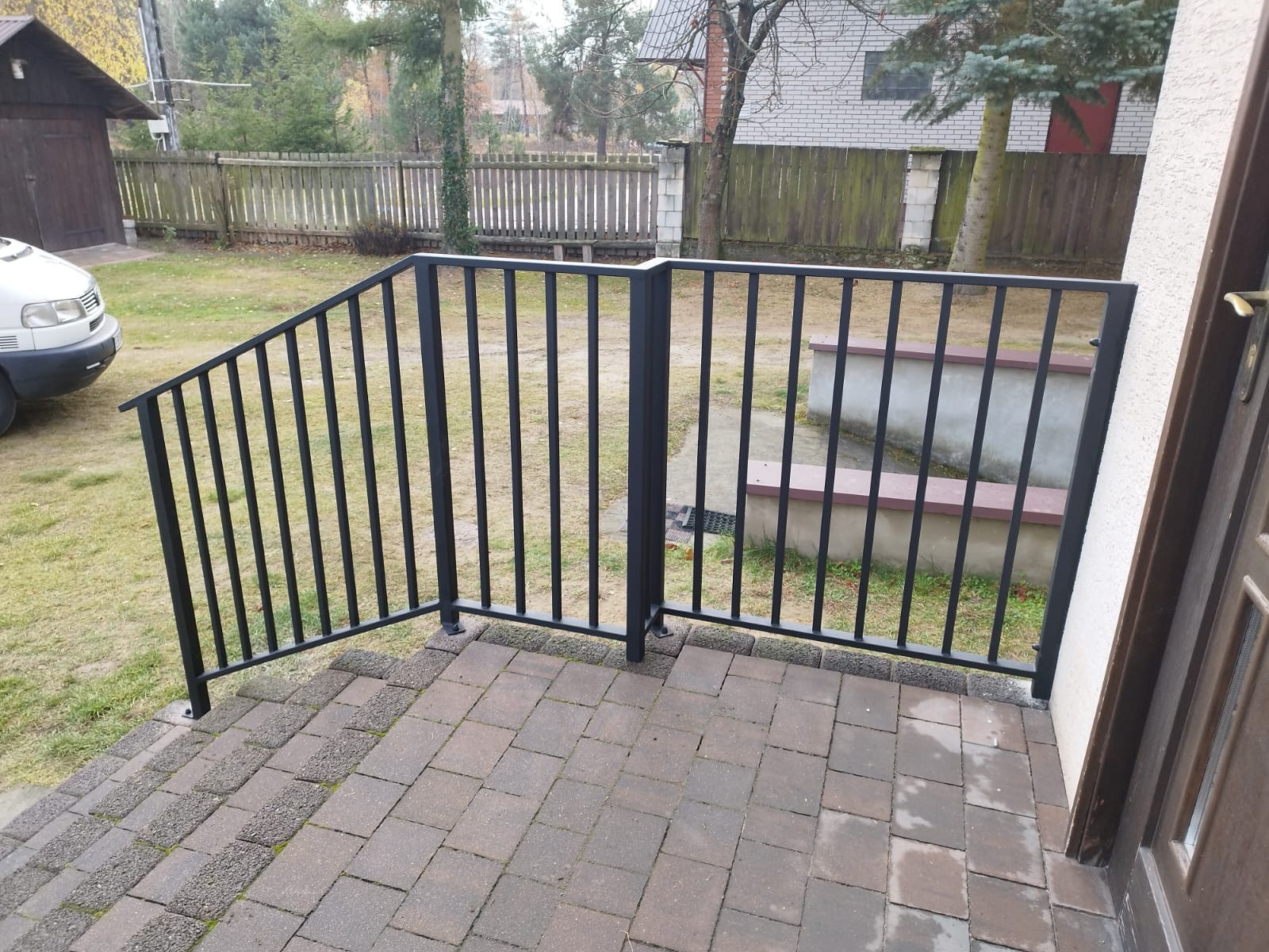 Czarne, metalowe barierki schodowe z pionowymi szczeblami, prowadzące do drzwi wejściowych, z kostki brukowej na podłożu, widok na trawnik i ogrodzenie w tle.