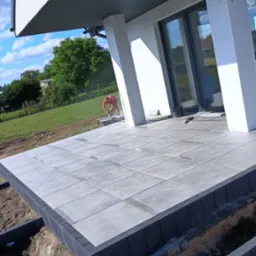 Układanie szarych płyt tarasowych z ciemną obwódką na zewnątrz domu. W tle brukarz przycina płytki. Widoczne narzędzia i przygotowane podłoże.