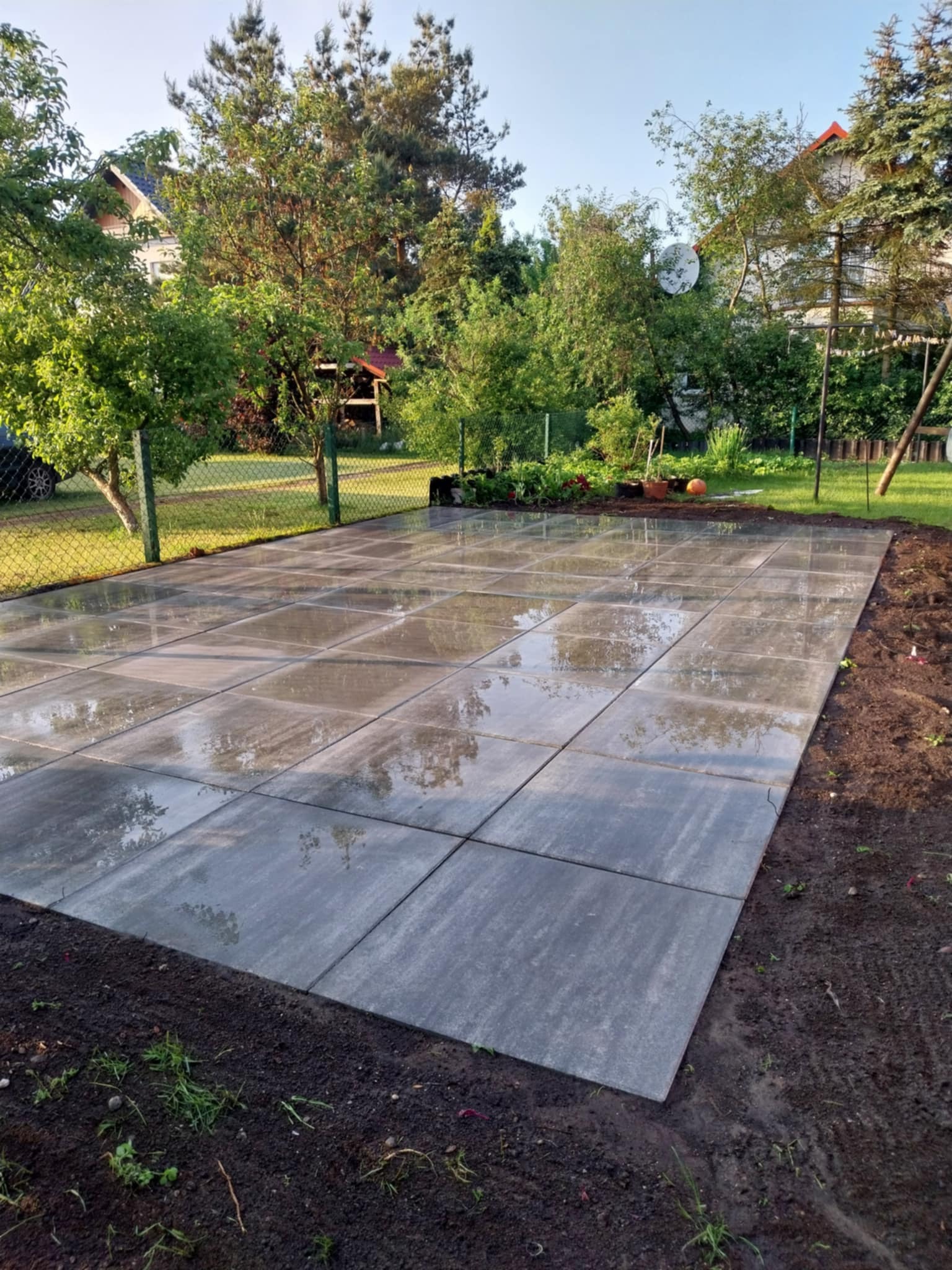 Świeżo ułożona, mokra, szara kostka brukowa na posesji, widok z góry, zarys domu w tle, trawa i drzewa. Kostka ułożona w kwadraty z widocznymi refleksami.
