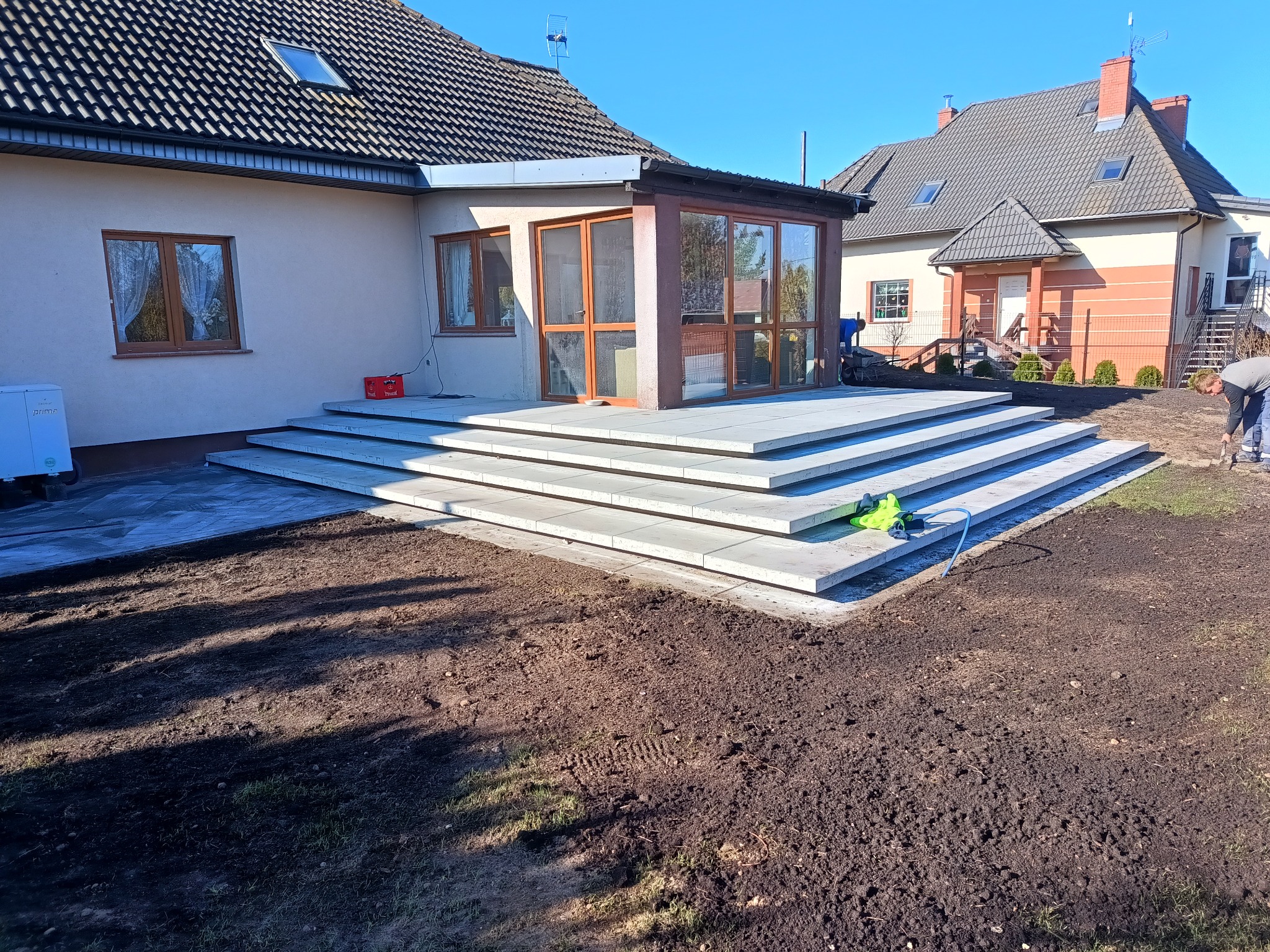 Nowoczesny taras z jasnoszarej kostki brukowej, schody prowadzące do domu z oranżerią, świeżo przygotowany teren pod trawnik, praca w toku.