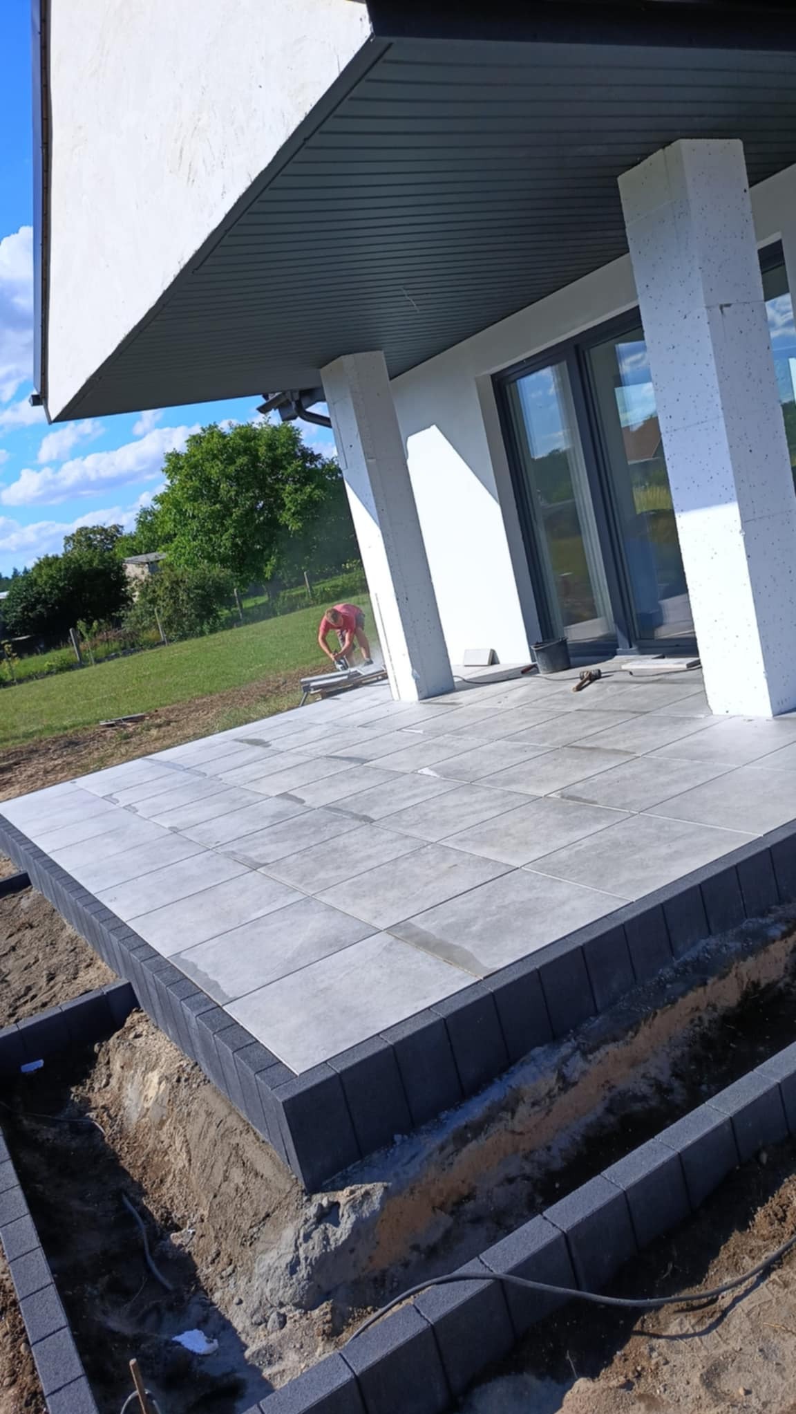 Układanie szarych płyt tarasowych z ciemną obwódką na zewnątrz domu. W tle brukarz przycina płytki. Widoczne narzędzia i przygotowane podłoże.