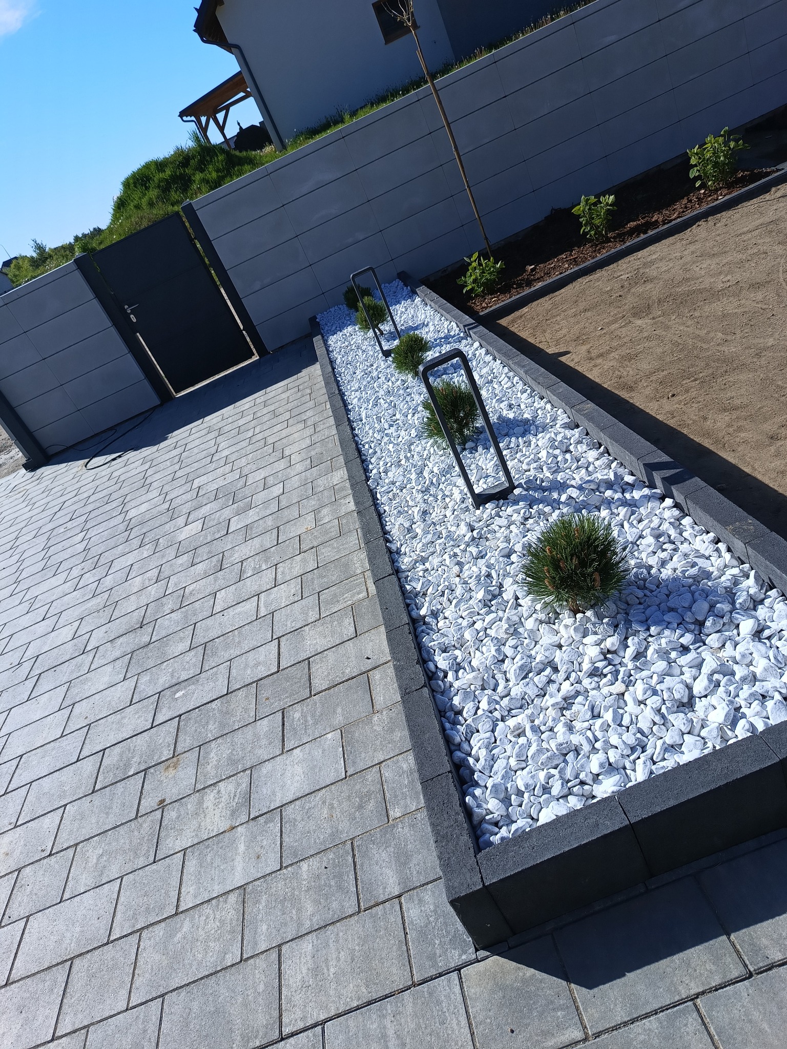 Szara kostka brukowa na podjeździe, obok rabata z białymi kamieniami, niskimi krzewami i nowoczesnymi lampami. W tle szary mur i błękitne niebo.