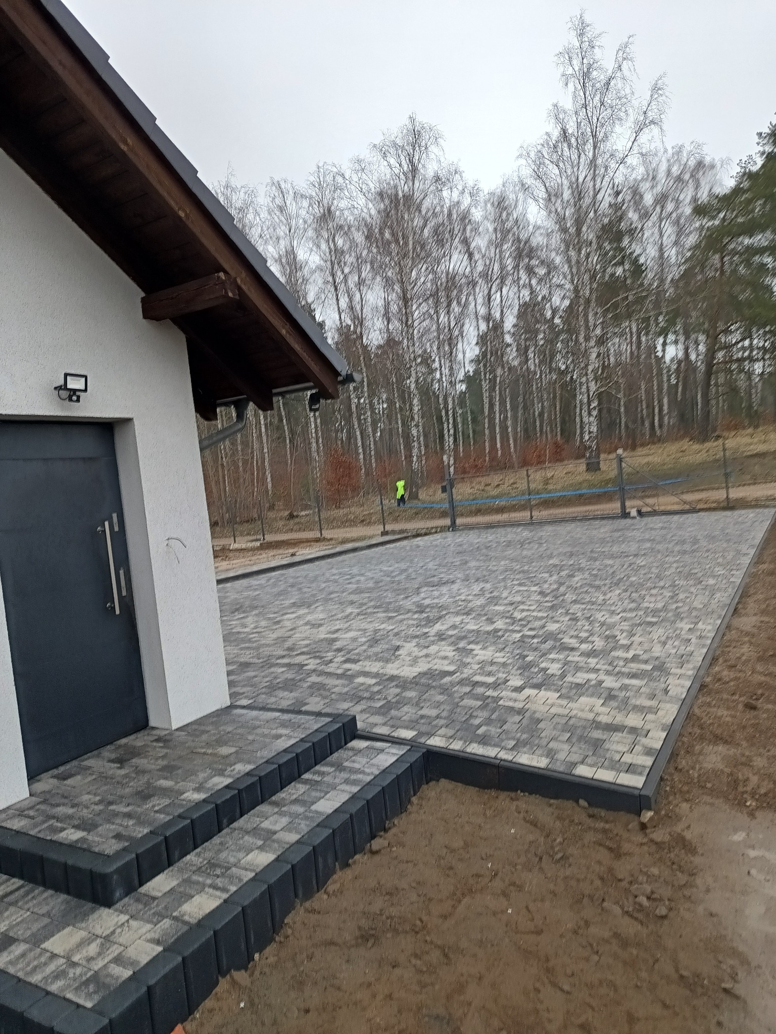 Nowoczesny podjazd z kostki brukowej w odcieniach szarości, z obrzeżami w kolorze grafitowym. Schody z kostki prowadzą do drzwi wejściowych domu.