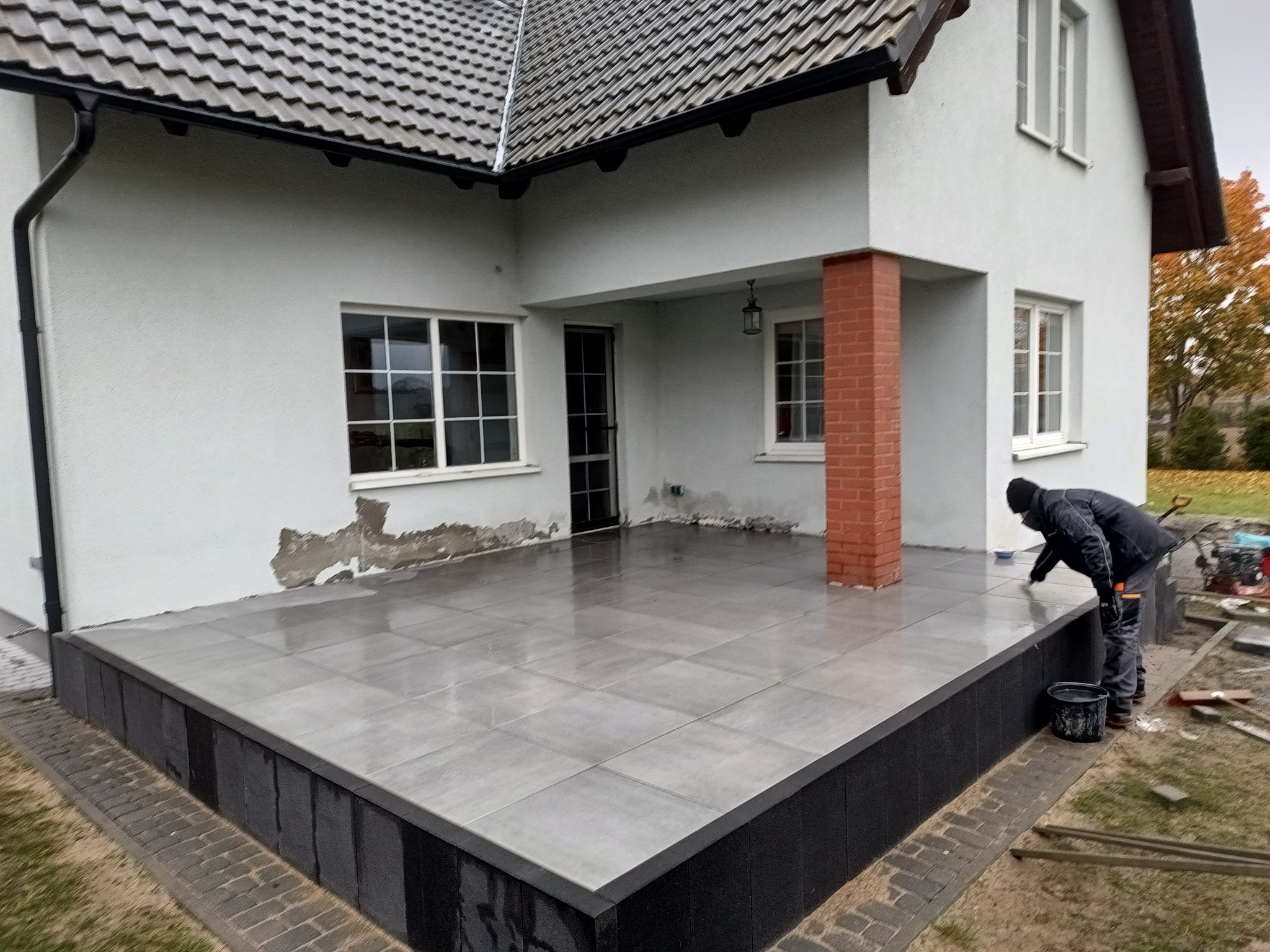 Szara, gładka, ceramiczna podłoga tarasu przy białym domu. Osoba czyści powierzchnię. Widoczne narzędzia i wiadro. Estetyczne wykończenie.