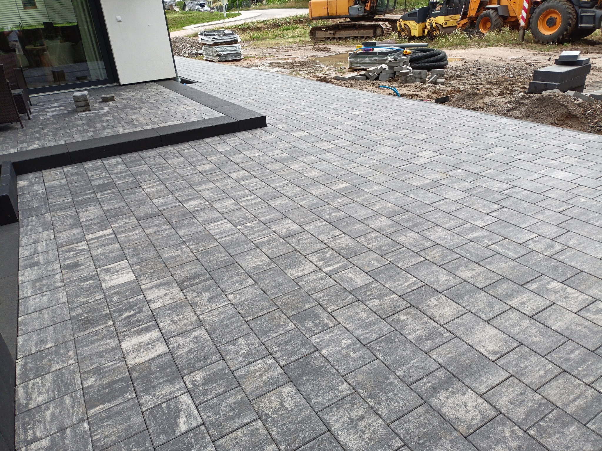 Ułożona szara kostka brukowa na podjeździe z czarnym obrzeżem. W tle maszyny budowlane i stosy kostki, sugerujące trwające prace wykończeniowe.