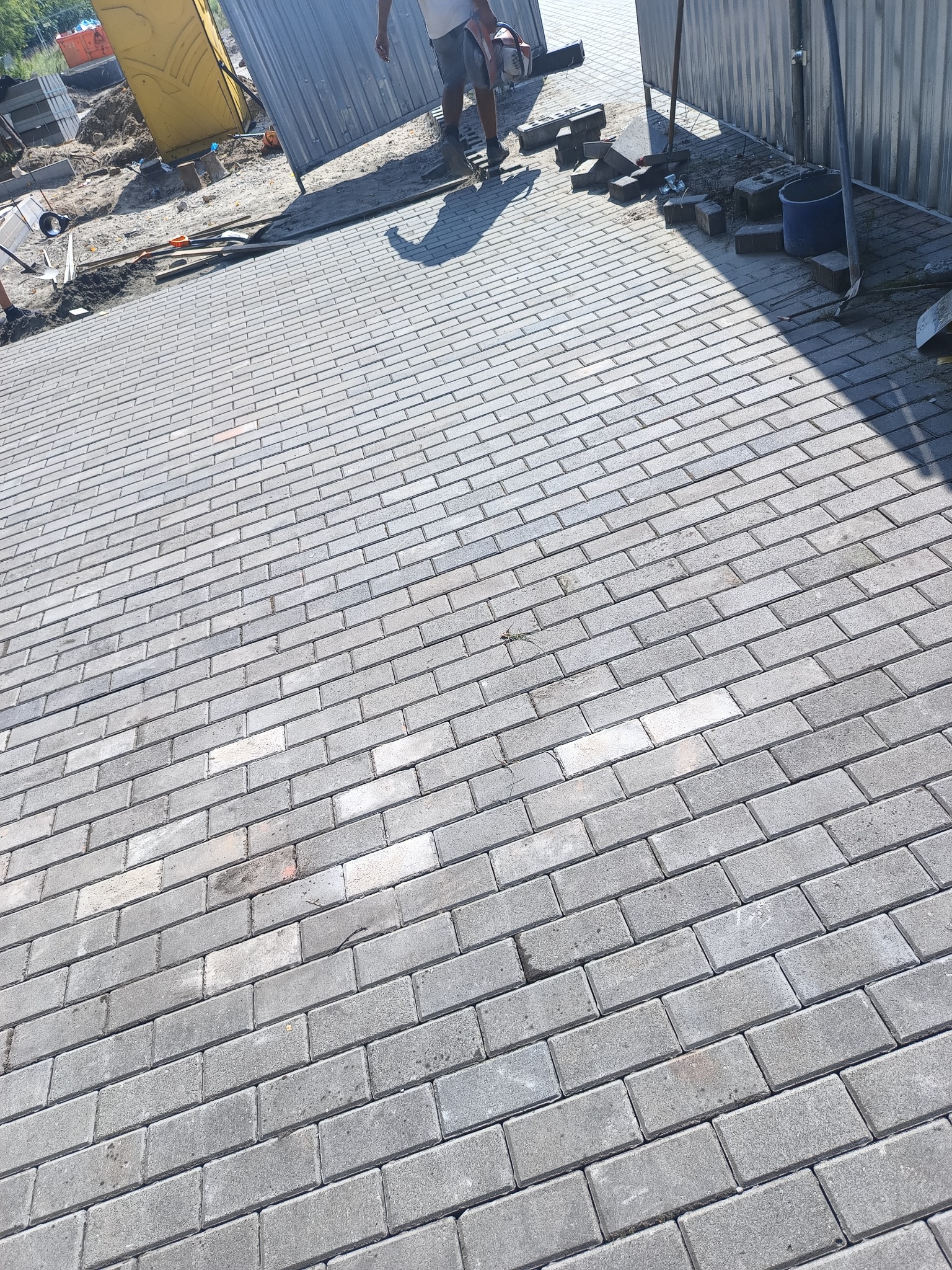 Fragment ułożonej, szarej kostki brukowej. W tle widoczny pracownik z przecinarką do kostki i tymczasowe ogrodzenie.