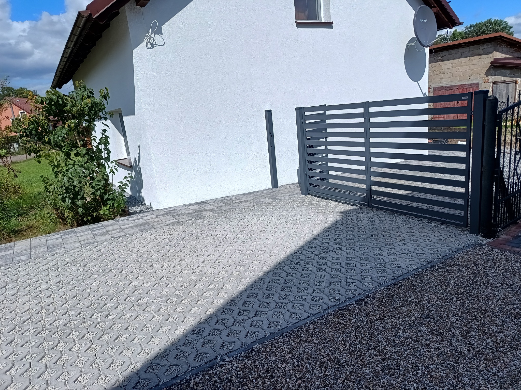Szara ażurowa kostka brukowa wypełniona drobnym kamieniem, prowadząca do nowoczesnej, poziomej bramy wjazdowej na tle białego domu z brązowym dachem.