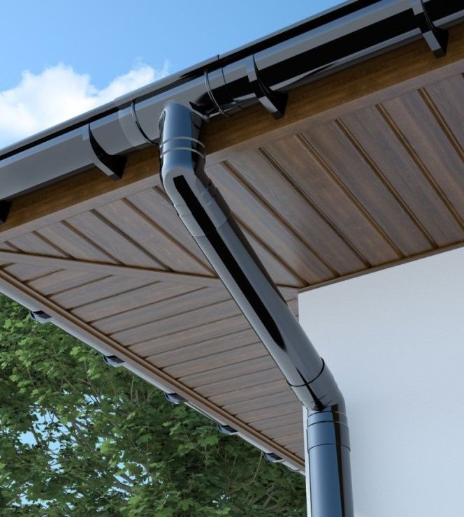 Błyszcząca, czarna rynna spustowa zamontowana na brązowej podbitce dachowej i białej ścianie budynku, widok z dołu na tle błękitnego nieba i zieleni drzew.