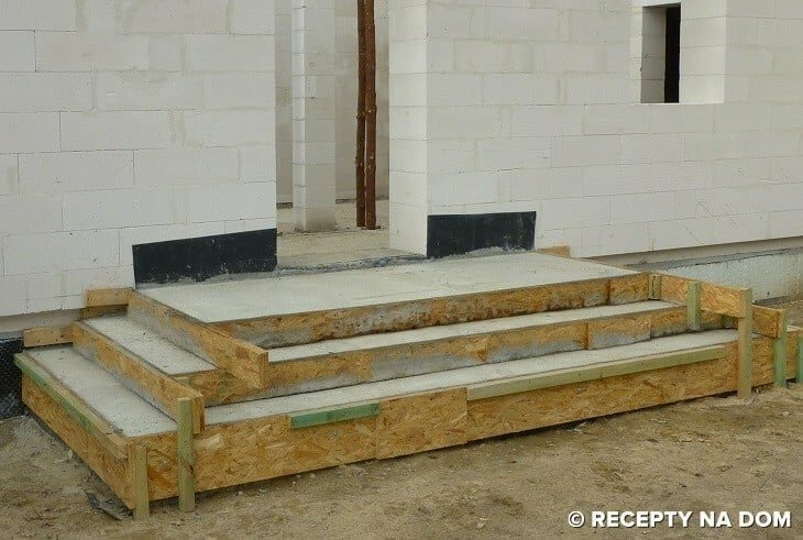Betonowe schody wejściowe w trakcie budowy, z drewnianym szalunkiem, na tle ściany z betonu komórkowego i przygotowanego wejścia do budynku.