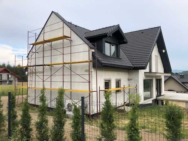 Dom jednorodzinny w trakcie ocieplania elewacji, widoczne rusztowania przy ścianie szczytowej i bocznej, czarne dachówki i częściowo otynkowana fasada.