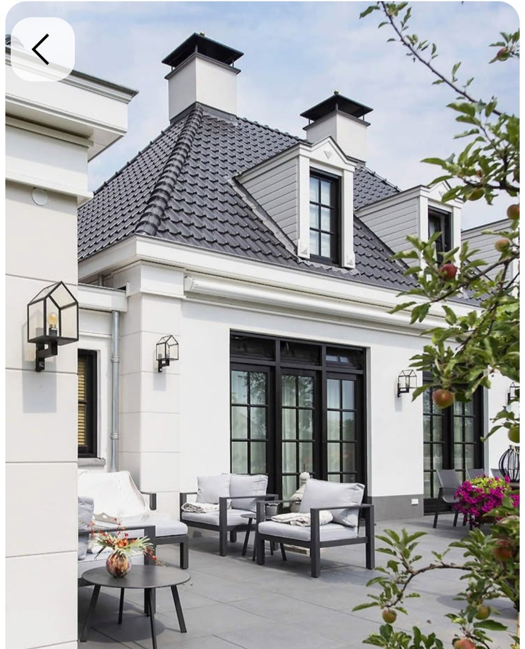 Biały dom z czarnym dachem i oknami, widok z tarasu z szarymi meblami ogrodowymi i jabłonią w tle.