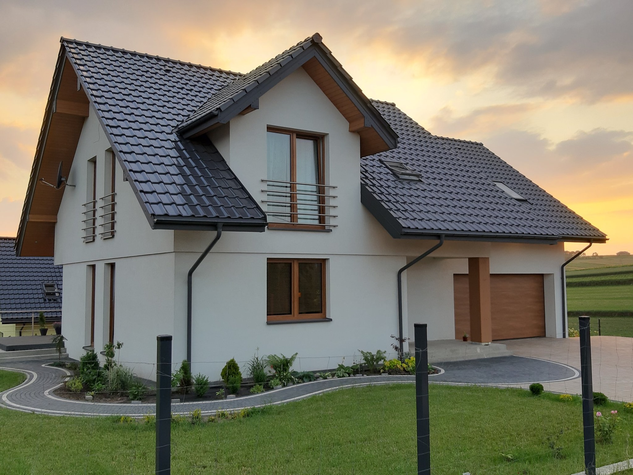 Nowoczesny dom jednorodzinny z szarą dachówką i jasną elewacją o zachodzie słońca, widoczne okna z drewnianą stolarką, zadbany trawnik i ścieżka z kostki brukowej.