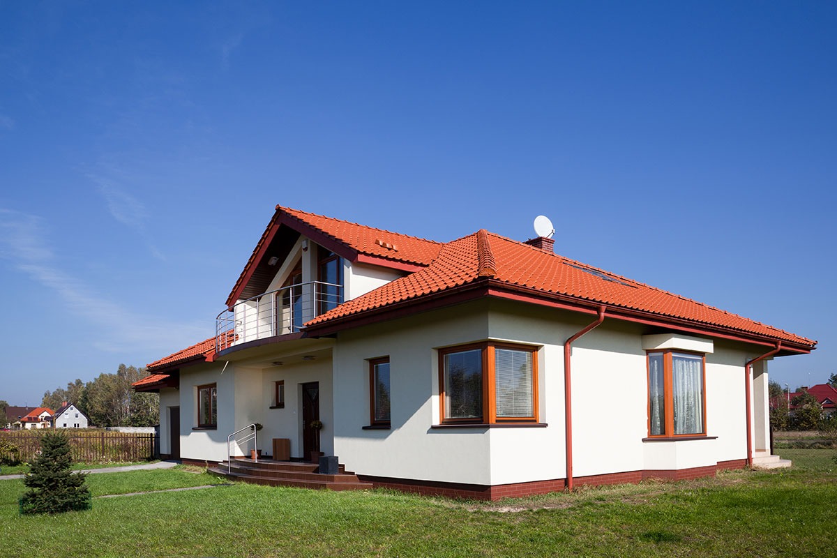 Dwupiętrowy dom jednorodzinny z czerwoną dachówką i białymi ścianami na tle błękitnego nieba, widoczna antena satelitarna na dachu.