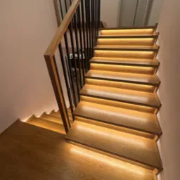 Drewniane schody z podświetleniem LED, czarne metalowe barierki. Widok z dołu, ukazujący detale drewna i ciepłe światło. Nowoczesny design.