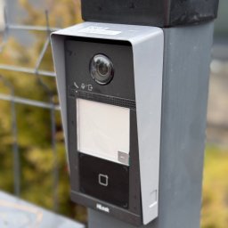 Smart Home Martin Marcin Rutkowski - Zamontowany wideodomofon HikLook na szarym słupku, z widoczną kamerą, ekranem i czytnikiem zbliżeniowym. W tle siatka ogrodzeniowa i zieleń.