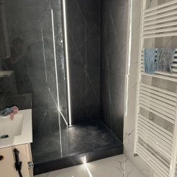 Smart Home Martin Marcin Rutkowski - Nowoczesna kabina prysznicowa z czarnymi płytkami imitującymi marmur, podświetlana pionowymi listwami LED, z grzejnikiem drabinkowym i marmurową podłogą.