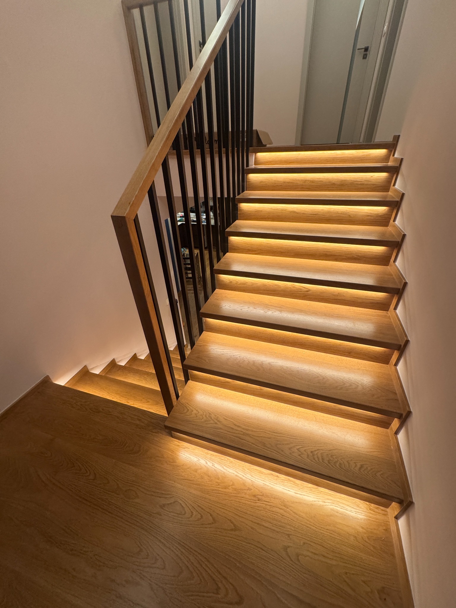 Drewniane schody z podświetleniem LED, czarne metalowe barierki. Widok z dołu, ukazujący detale drewna i ciepłe światło. Nowoczesny design.