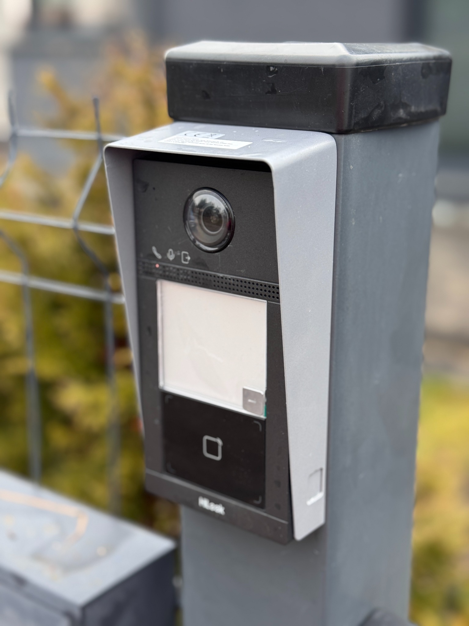 Zamontowany wideodomofon HikLook na szarym słupku, z widoczną kamerą, ekranem i czytnikiem zbliżeniowym. W tle siatka ogrodzeniowa i zieleń.