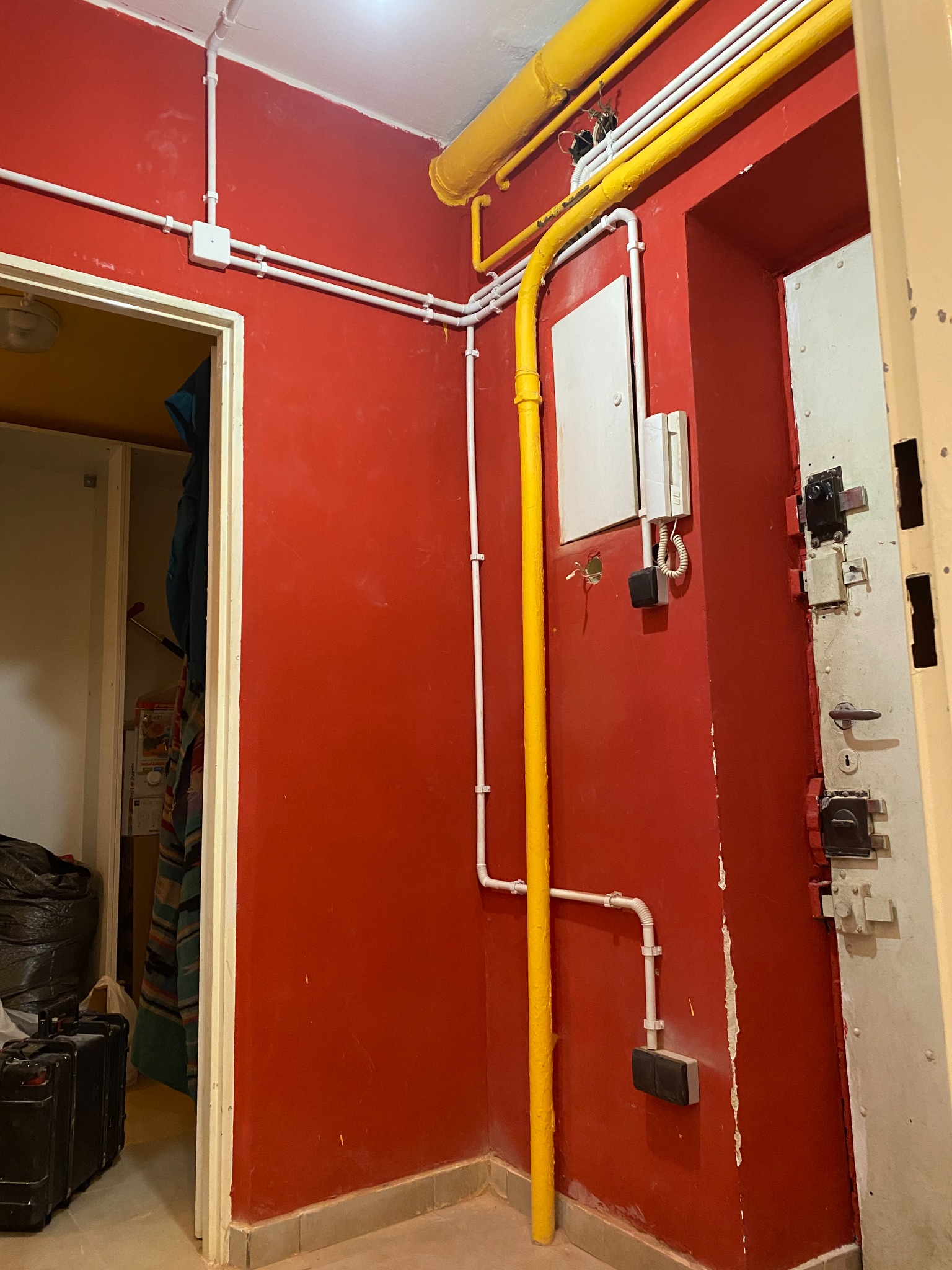 Czerwone wnętrze z instalacjami: rury białe i żółte, puszki elektryczne, domofon przy masywnych, metalowych drzwiach. Widoczne elementy instalacji.