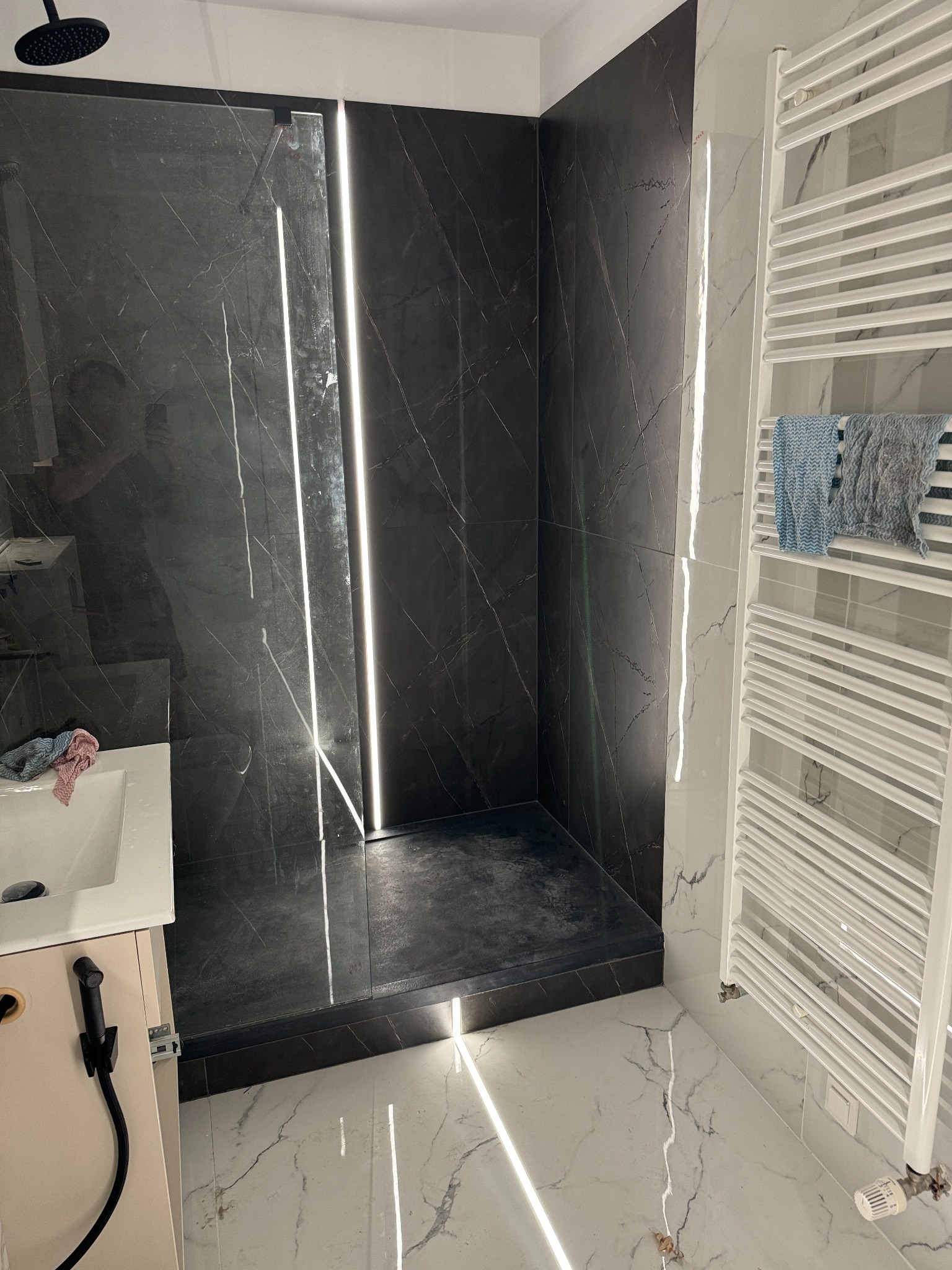 Nowoczesna kabina prysznicowa z czarnymi płytkami imitującymi marmur, podświetlana pionowymi listwami LED, z grzejnikiem drabinkowym i marmurową podłogą.
