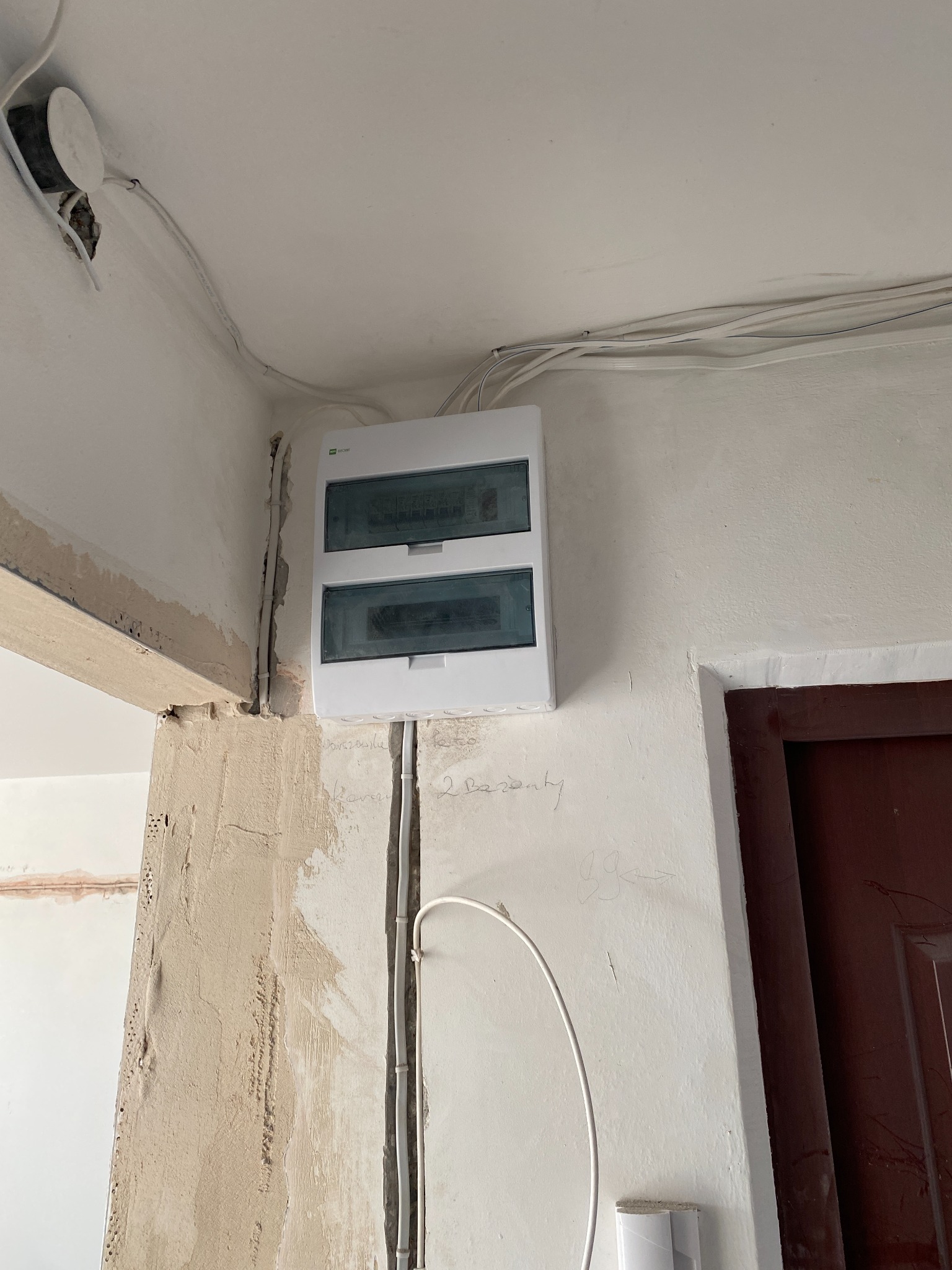 Biała skrzynka rozdzielcza z widocznymi bezpiecznikami, zamontowana na ścianie w trakcie remontu. Widoczne przewody i surowy tynk.