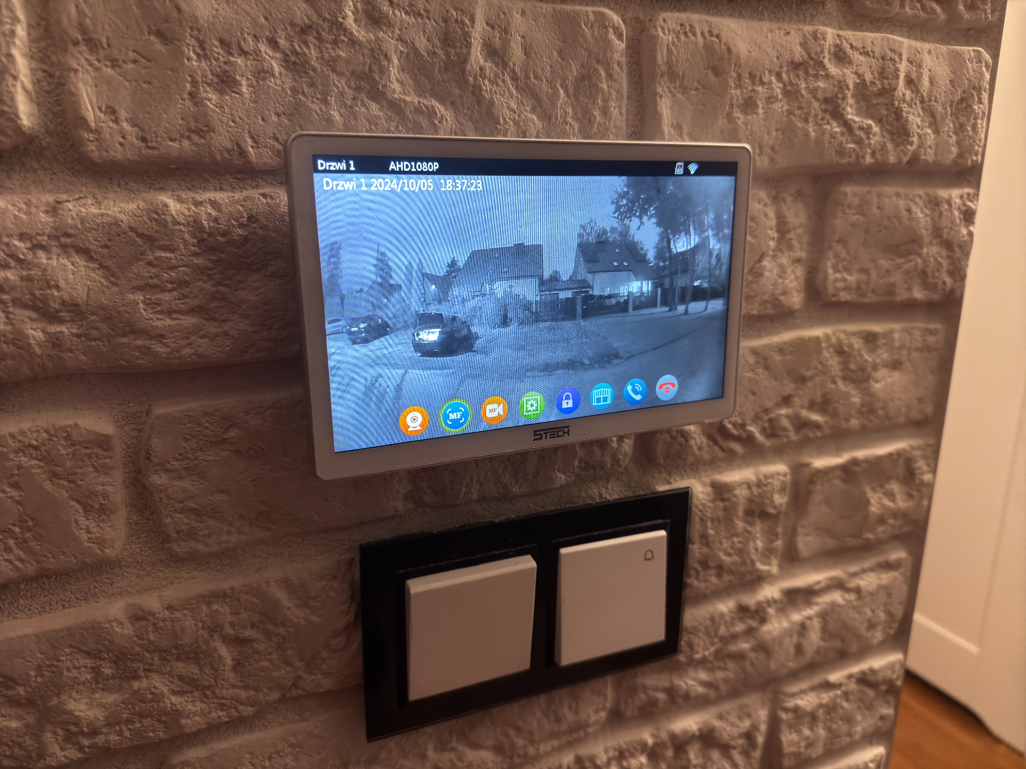 Zamontowany na ceglanej ścianie monitor systemu monitoringu z widokiem na ulicę, obok włącznik światła. Ekran pokazuje obraz z kamery AHD1080P.