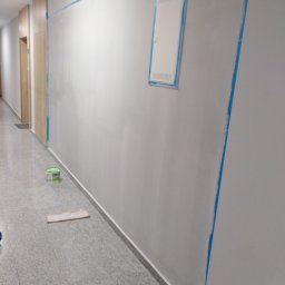 Zbigniew Grożyński Gro-Bud Usług remontowo budowlane - Świeżo pomalowany korytarz w Gdańsku z zabezpieczonymi listwami i framugami taśmą malarską, widoczny otwarty pojemnik z farbą i narzędzie malarskie na podłodze.
